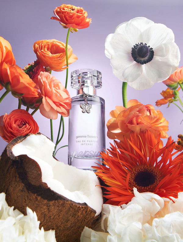 Gimmie Flowers Eau De Parfum Intense 1.7 fl oz bottle surrounded by colorful flowers and coconuts on a light purple background