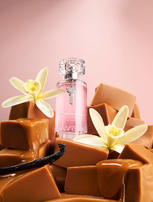 Caramel Temptress Eau De Parfum Intense 1.7 fl oz perfume bottle with vanilla flowers and caramel pieces on a pink background